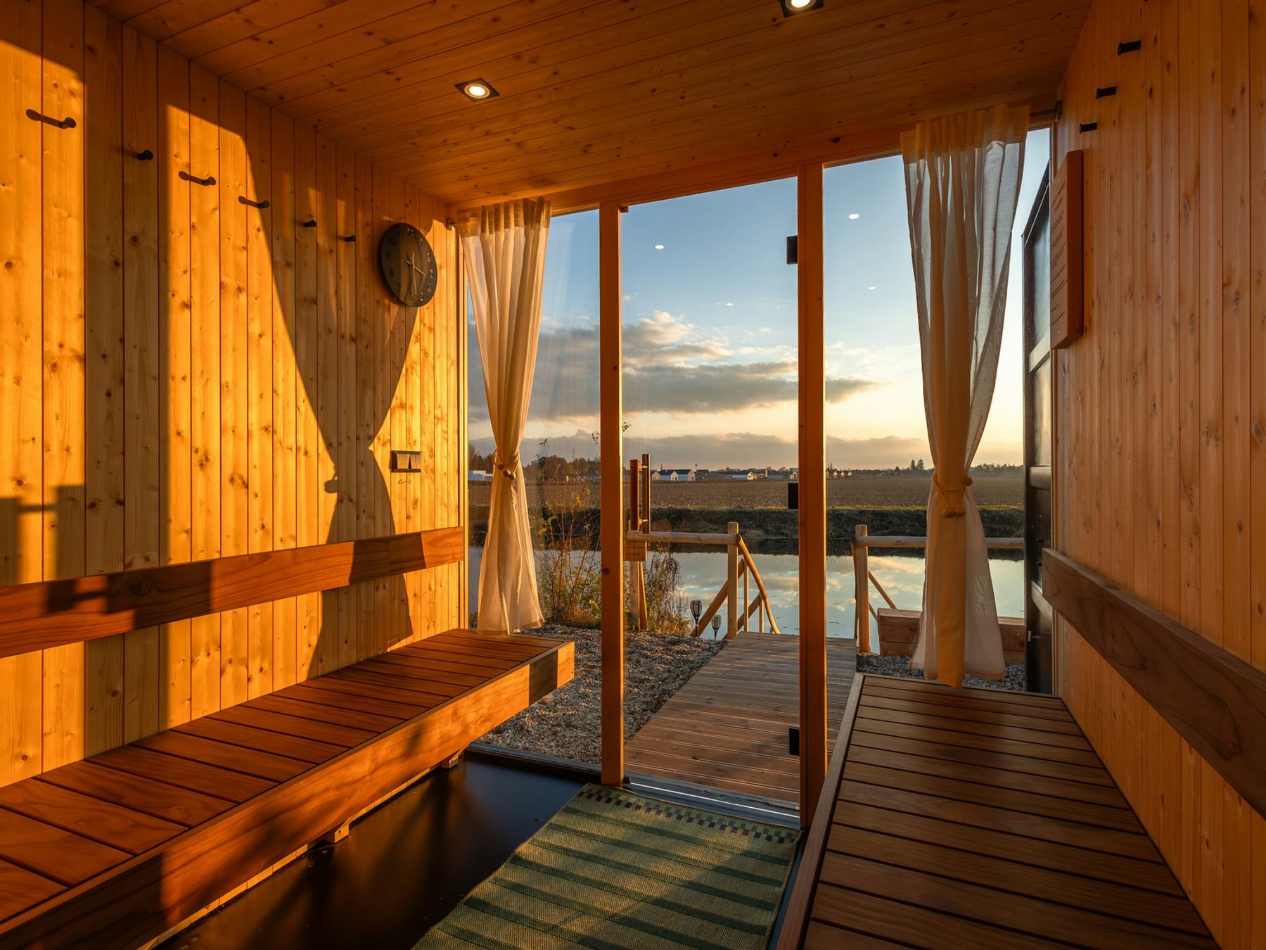 A light wooden sauna with its glass door open to the jetty at sunset